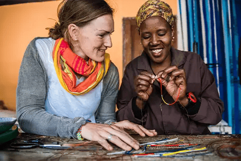 Two women sitting at a table smiling as they work together on colorful beaded jewelry. One woman threads beads while the other watches and helps arrange materials