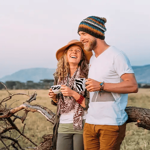 A couple laughing and enjoying a 'sundowner' experience in Serengeti National Park. They are holding zebra-striped mugs in the golden light of sunset, dressed in warm, casual safari gear.